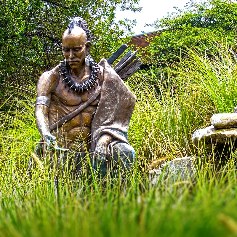 Bronze statue of a hunter with a bow and quiver, kneeling among tall grass in a garden.