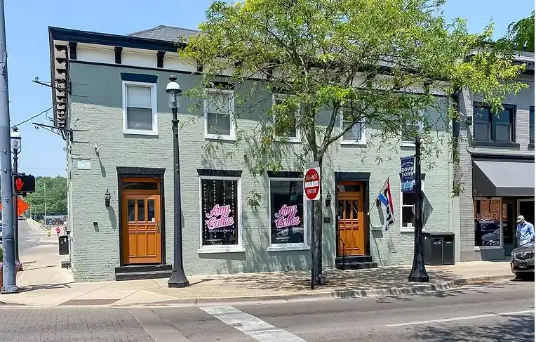 Two-story green brick corner Amy Cakes storefront with wooden doors and display windows featuring tasty treats inside, flanked by trees on a sunny urban street.