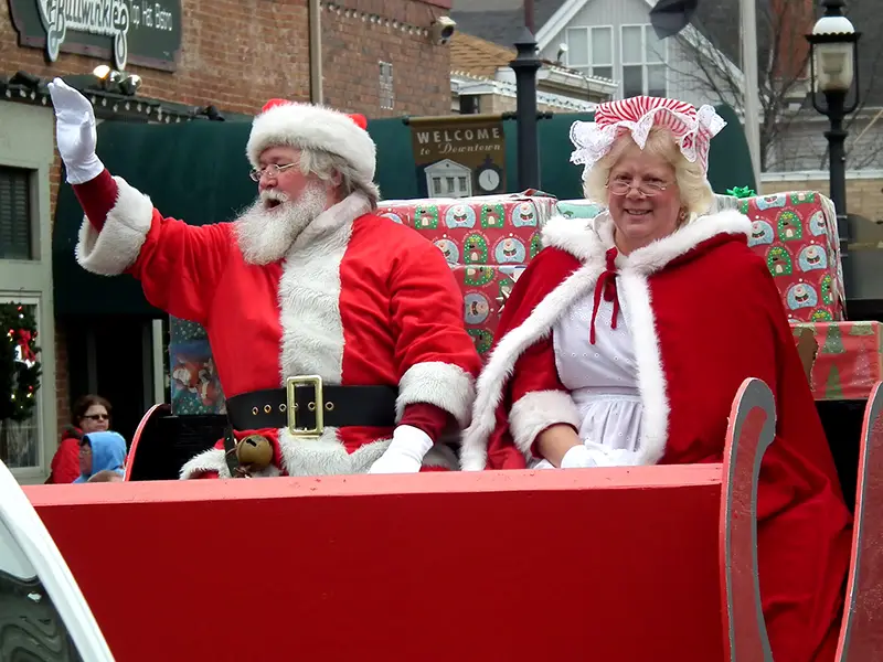 Santa Claus waves from a red sleigh with a smiling woman in a festive outfit during a Christmas parade.