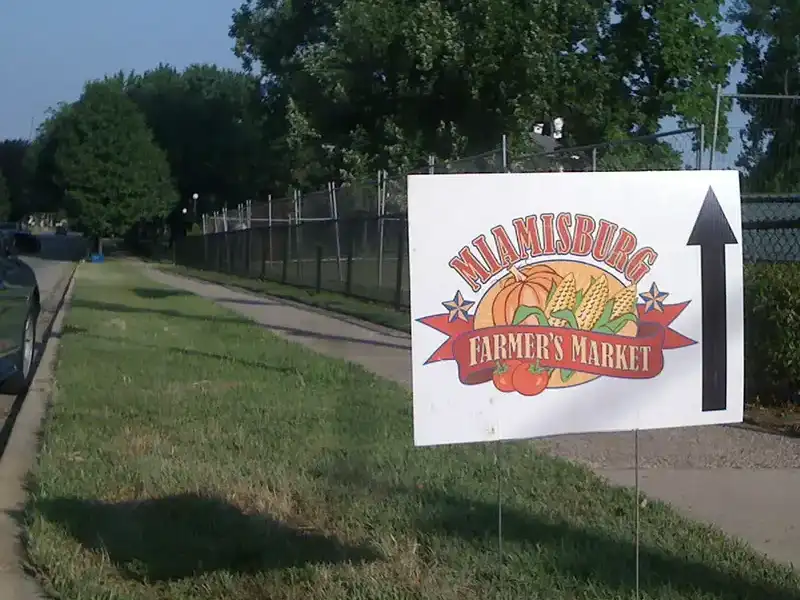A white sign with a colorful farmers market logo and a bold upward arrow stands beside a sidewalk on a grassy verge.