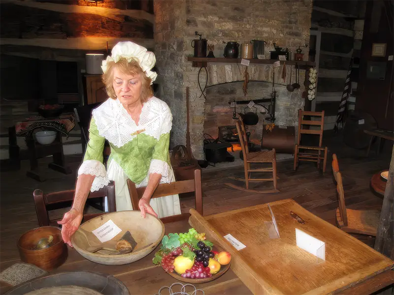 A woman in a white bonnet and green period dress stands behind a wooden table with a basket of fruit in a rustic, old-fashioned kitchen.