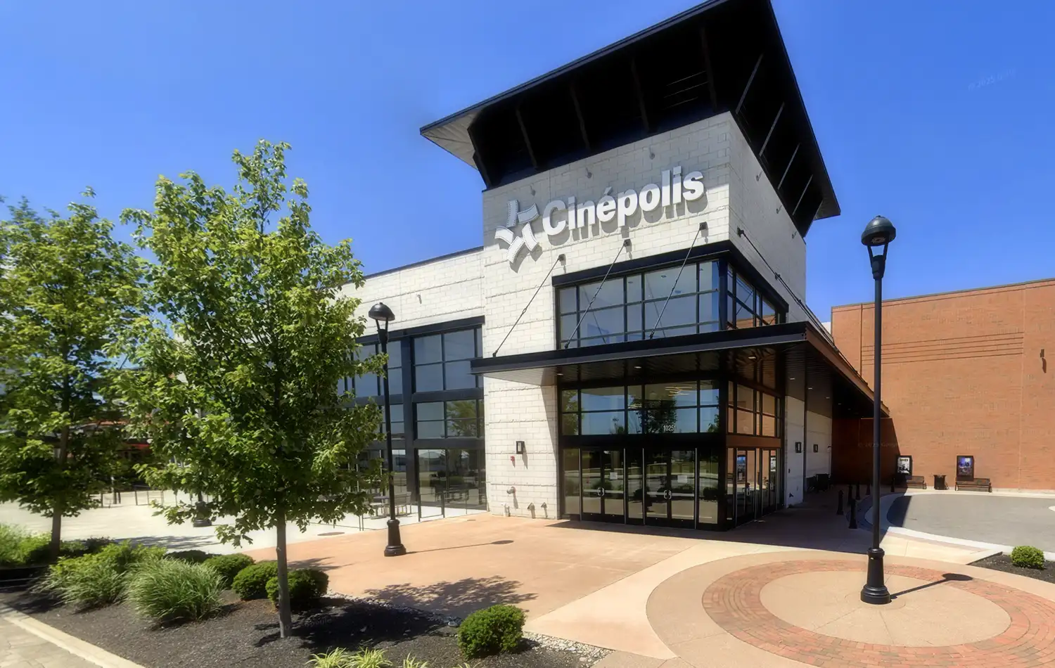 Modern two-story commercial building with a glass storefront and a tall dark roof overhang, surrounded by trees and a circular brick-paved plaza.