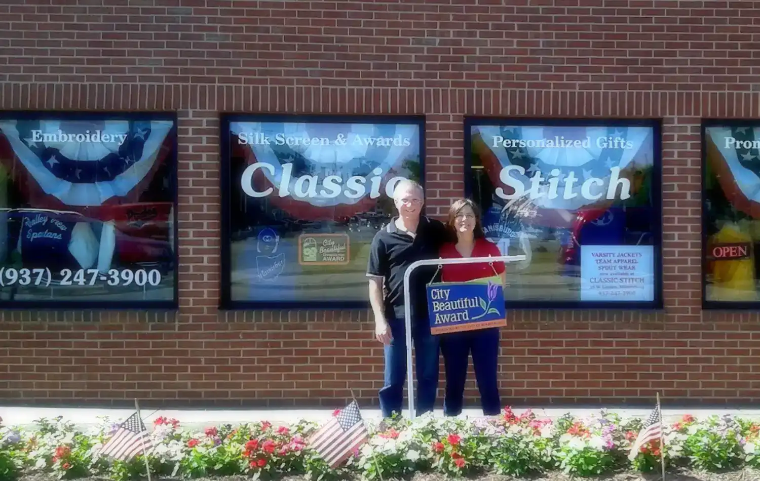 Classic Stitch Custom Apparel Two people stand outside a brick storefront with window signs reading “Classic Stitch” and flowers in the foreground.