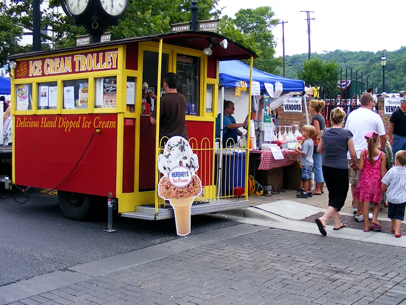 A red and yellow ice cream trolley serving customers at an outdoor event, with people lined up nearby.