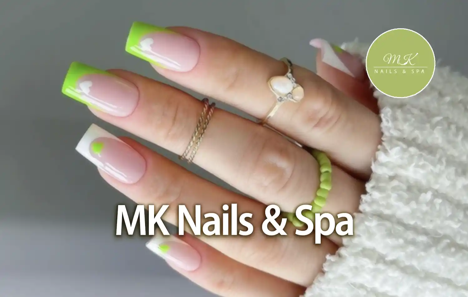 MK Nails and Spa Close-up of a hand with long nails featuring neon-green tips, wearing gold rings and a white knit sleeve.