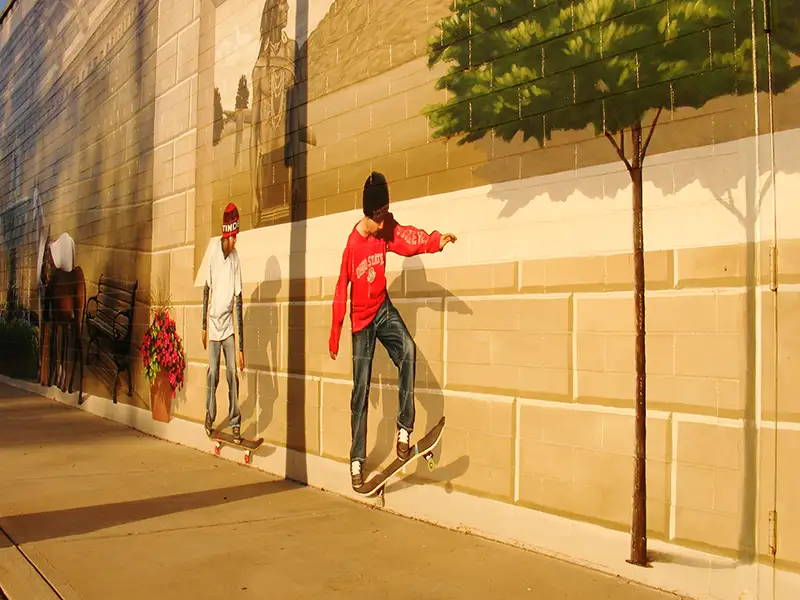 A skateboarder wearing a red shirt skates along a sunlit beige wall beside a sidewalk.