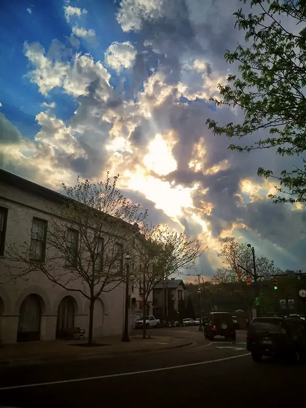A city street at dusk with an arched building on the left, leafless trees, cars on the road, and a dramatic sky with sunlight breaking through clouds.