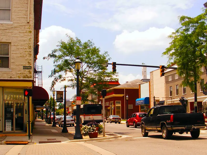 Sunny downtown street with brick storefronts, trees, traffic lights, and cars.