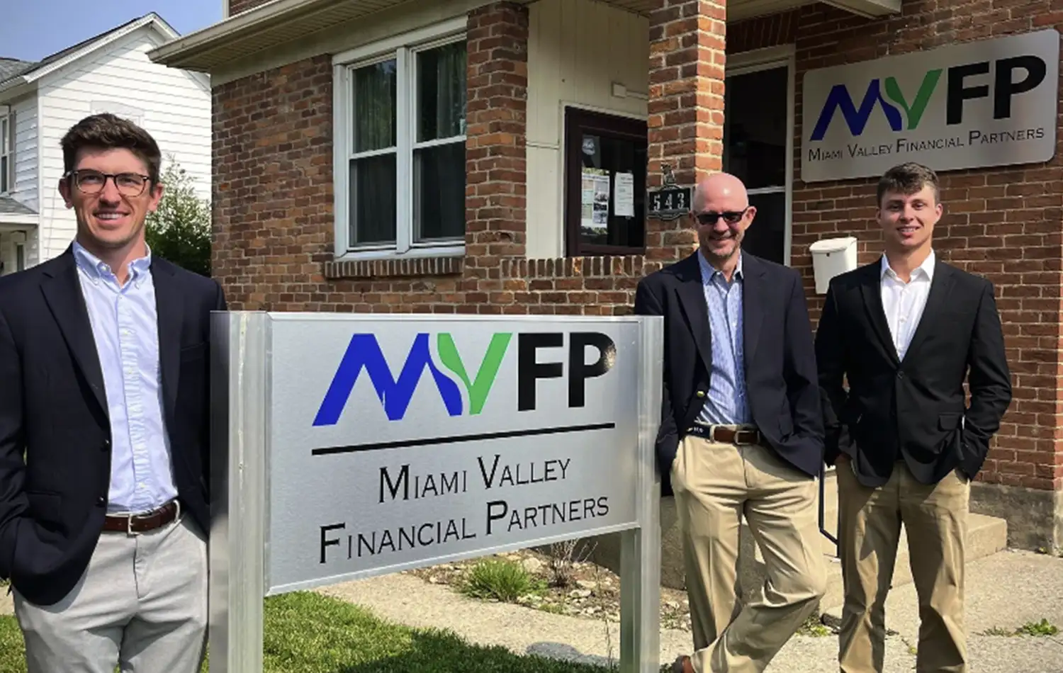 Three men in business attire pose outside a brick office building beside a Miami Valley Financial Partners sign.