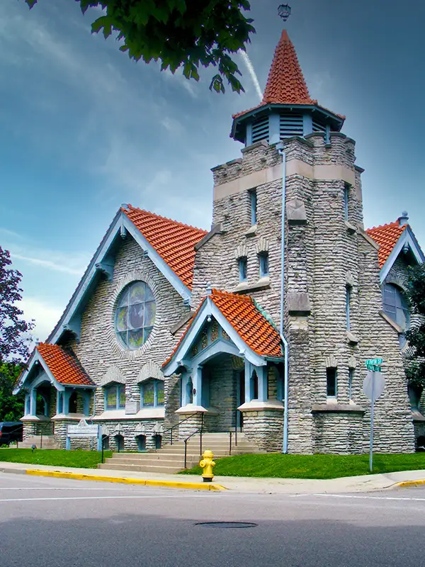 Stone church with a red-tiled roof, a tall conical tower, and a circular stained-glass window, set on a grassy area beside the street.