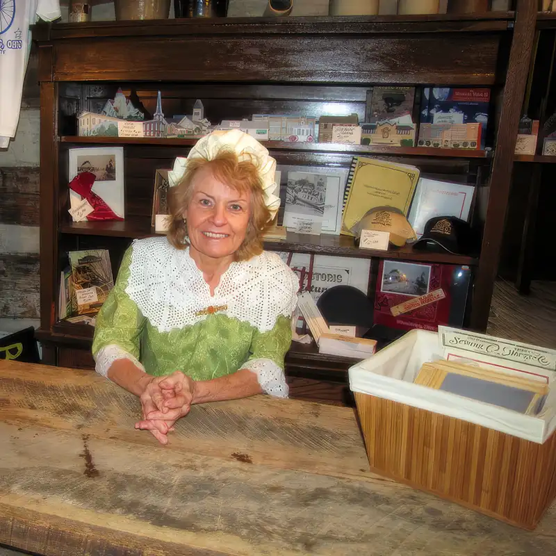 Smiling blonde woman in a green dress with a white lace collar sits behind a wooden counter, with shelves of books and framed items in the background.